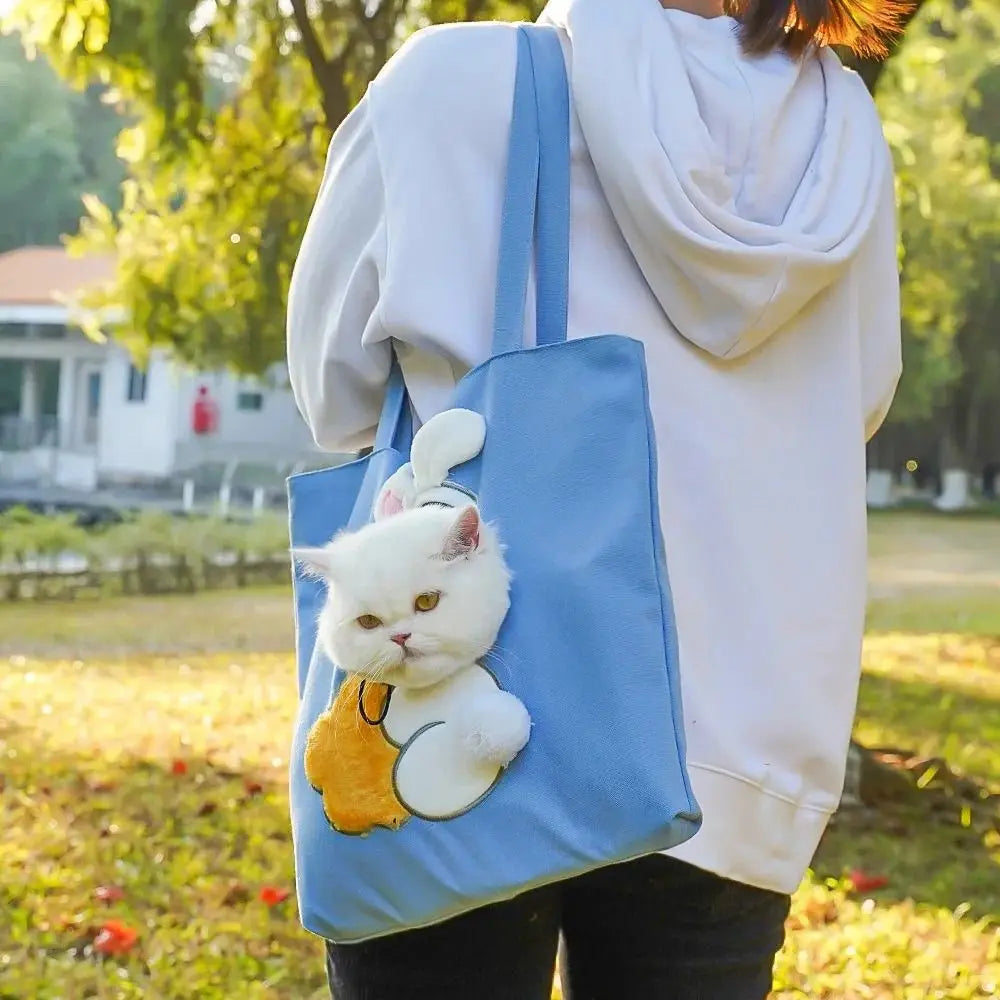 A person wearing a white hoodie carries a blue portable crossbody pet carrier bag featuring a white cat design outdoors.