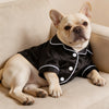 Un bulldog francés de color claro vistiendo una camisa de satén negra con ribetes blancos, sentado en un sofá beige.