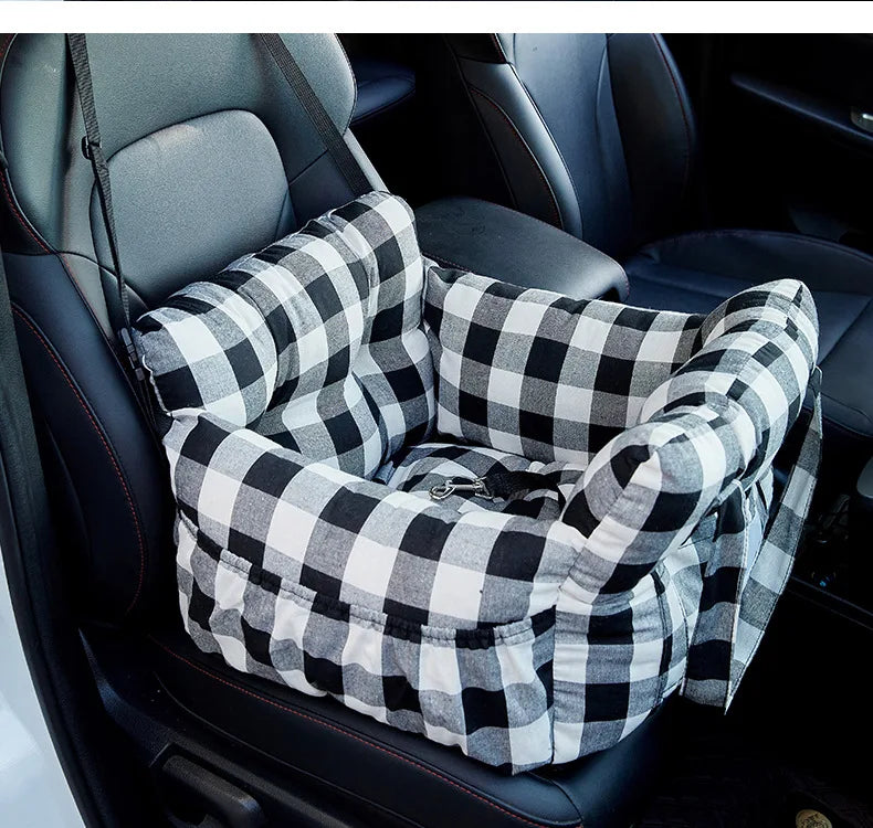A plaid pet car seat with safety straps is placed in a dark car interior.