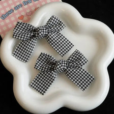 A pair of black and white plaid fabric bow hair clips displayed on a white ceramic dish.