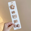 A hand holds a white card displaying five handmade flower-shaped baby hair clips in various shades of brown and beige.