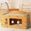 A light wood cat scratcher house and lounge bed with a padded interior and hanging balls.