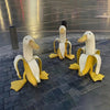 A set of cute resin banana duck garden sculptures in white and yellow sitting on a dark paved surface.