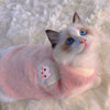 A fluffy white and blue cat wearing a pink embroidered vest on a white, furry surface.