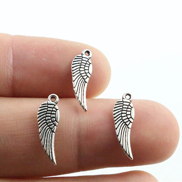 A hand holds several antique silver wing charms, showing their detail and size.