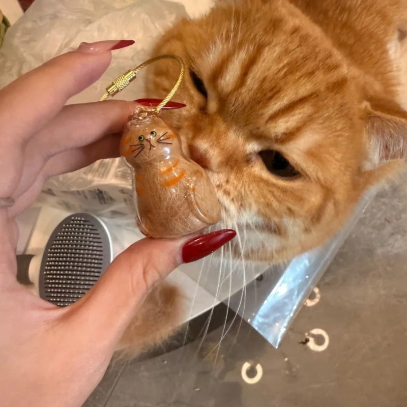 Eine Hand hält eine kleine, bernsteinfarbene Katzenfigur als Schlüsselanhänger mit goldener Schlaufe vor dem Gesicht einer or