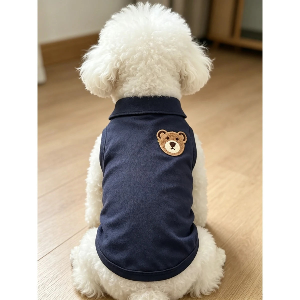 Un perro blanco peludo vistiendo un chaleco sin mangas azul marino con bordado de oso pardo en la espalda.