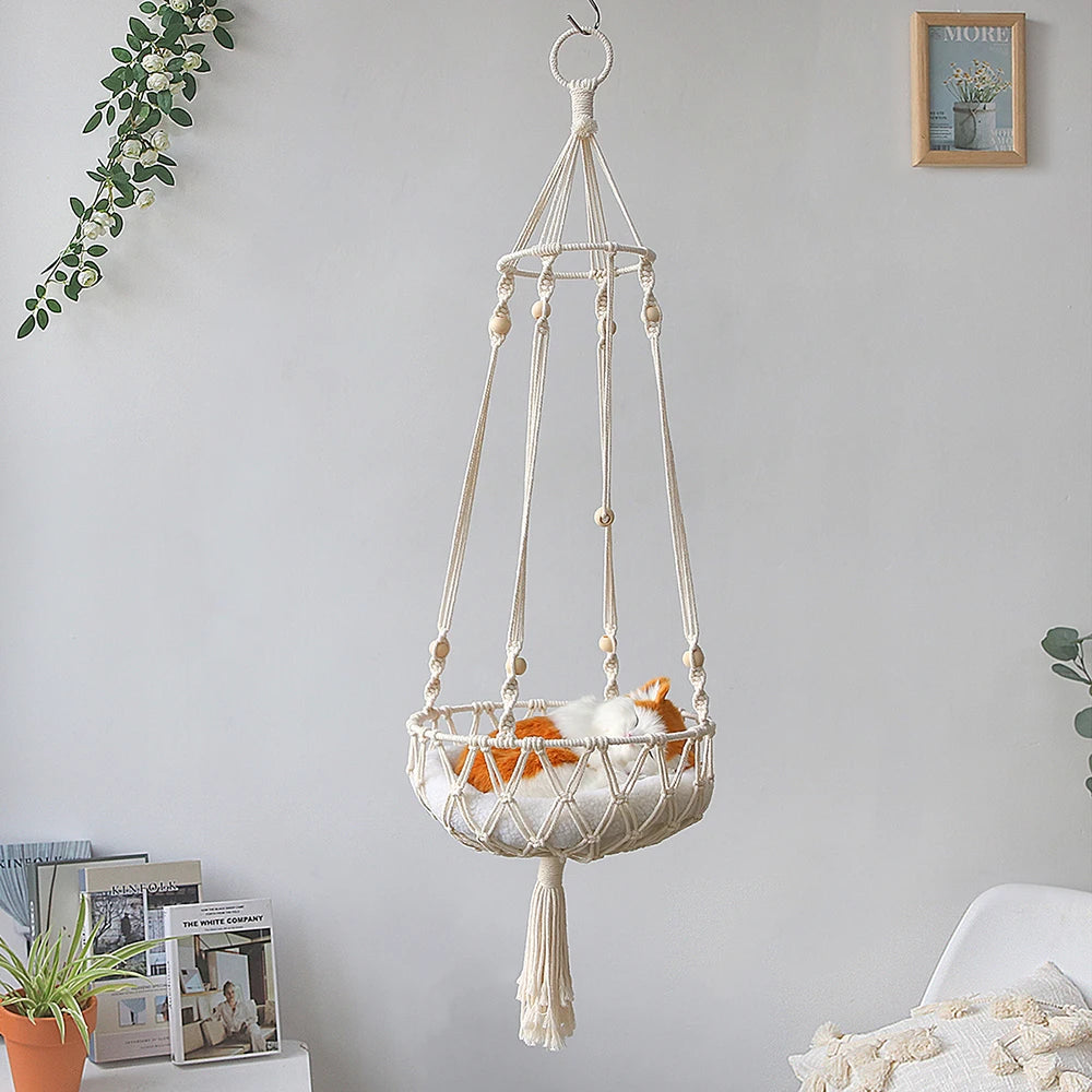 A bohemian macrame cat hammock bed for pet decor hangs on a wall, featuring a woven basket and a fluffy orange kitten.