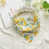A yellow and green floral paisley triangle head scarf is displayed on a white linen cloth next to a book.