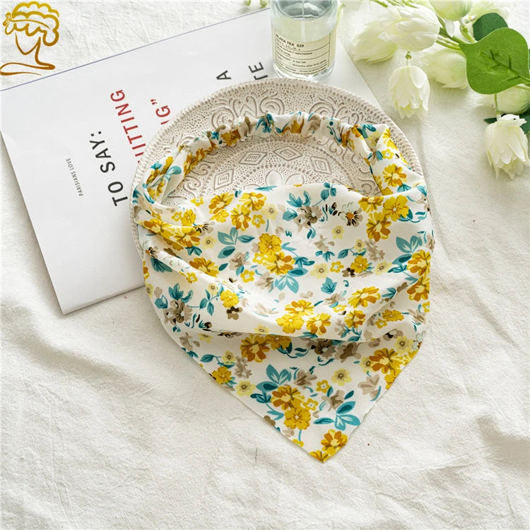 A yellow and green floral paisley triangle head scarf is displayed on a white linen cloth next to a book.