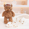 A brown teddy bear plush toy with a white crown, pajamas, and an eye mask displayed on a light surface.