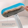 A hand is using a reusable dog hair brush with blue bristles and a gray silicone base to remove hair from a surface.