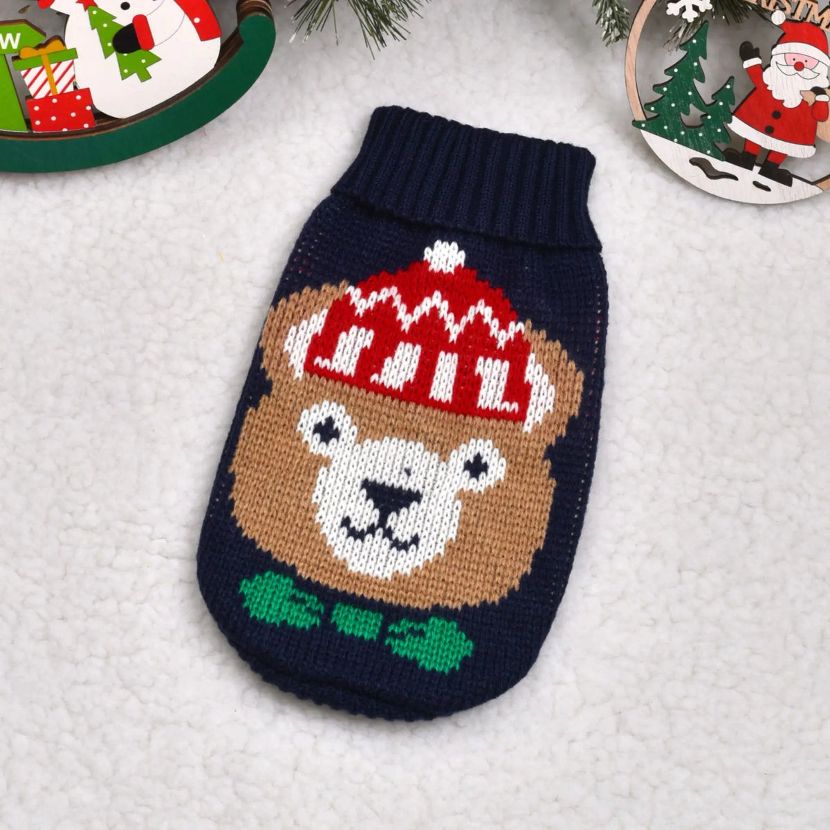 A knitted dog sweater featuring a brown bear face with a red hat and green collar, displayed on a white fuzzy surface.