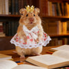 Ein flauschiger brauner Hamster mit goldener Krone und rosa Kleid mit Spitzenbesatz, sitzt auf Holzoberfläche neben Büchern.