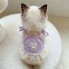 A small white cat wearing a lavender floral print dress with thin straps and a ruffled hem, sitting on a white fluffy surface