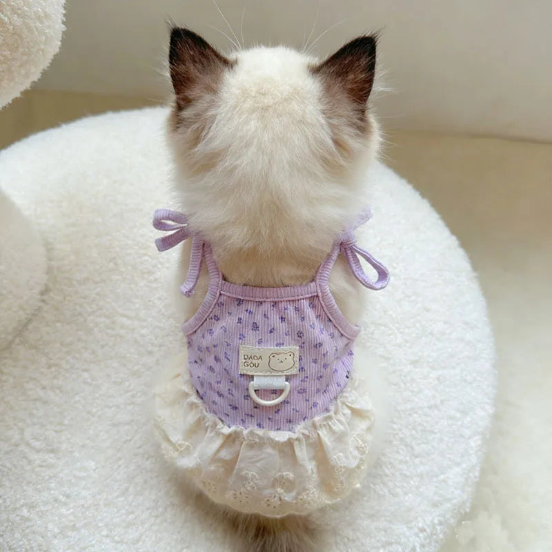 A small white cat wearing a lavender floral print dress with thin straps and a ruffled hem, sitting on a white fluffy surface
