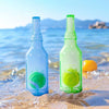 A clear glass bottle filled with bright green liquid and a floating green tennis ball on the sand near the ocean.