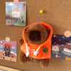 An orange capybara plush hoodie doll with small wings is displayed next to colorful cards on a brown surface.