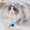 A fluffy gray and white cat is visible inside a mesh tunnel for indoor play with a colorful feather toy.