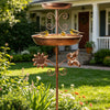 Baño de pájaros de bronce vintage montado en soporte con arabescos decorativos, en un jardín.