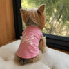 Un chien brun clair est allongé, regardant sur son dos, portant un débardeur rose avec du texte blanc.