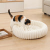 A tabby cat is resting in a round, white, plush pet bed with a bolster support on a light wooden floor.