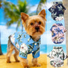 A golden-colored dog wearing a blue Hawaiian style summer beach shirt with floral and palm leaf patterns.