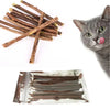 Dried natural herbal cat chew sticks arranged diagonally next to a gray cat with its tongue out.