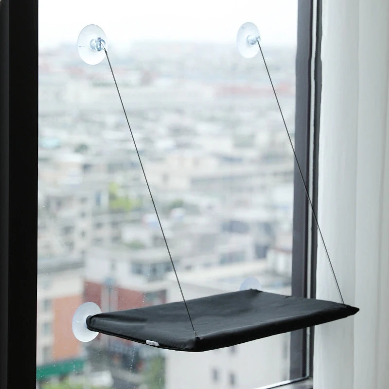 A black hanging hammock suspended by thin lines from suction cups, visible through a window overlooking a cityscape.