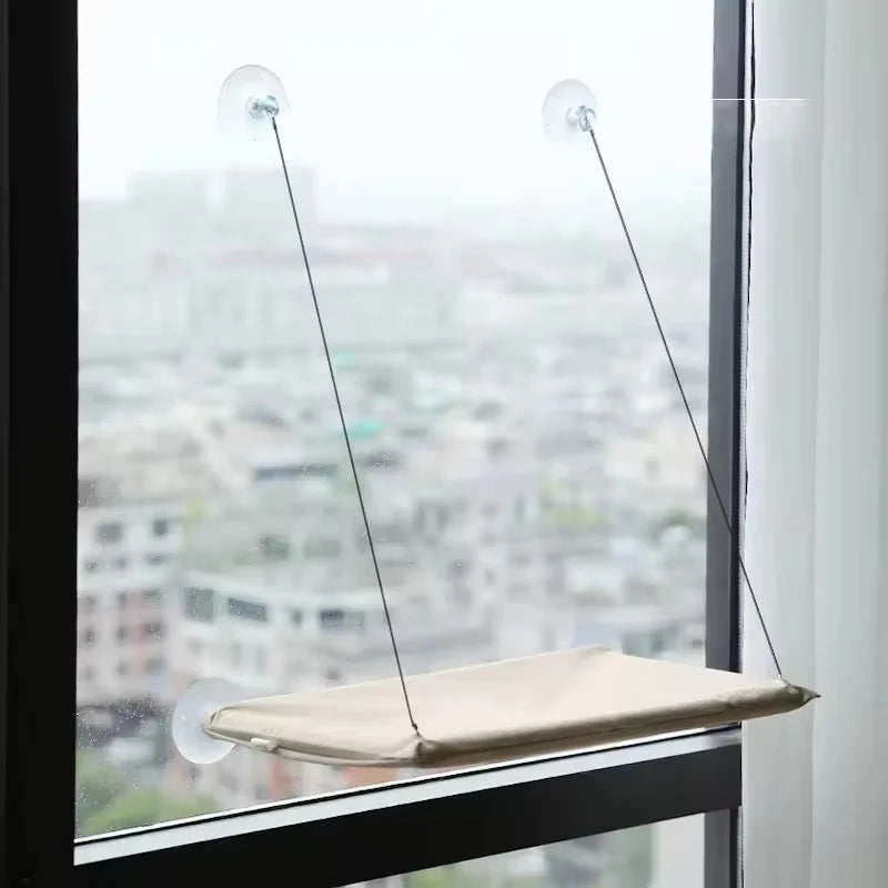 A beige hammock bed suspended by wires in a window, designed for a cat.