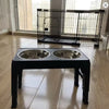 A black adjustable raised dog feeder with two stainless steel bowls sits on a light wooden floor.