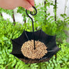 A black umbrella-shaped hanging bird feeder filled with seeds is being held by a hand in a green outdoor setting.