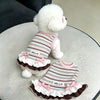 A white poodle wearing a striped dress with pink trim, bowknot, and ruffle on a light surface.