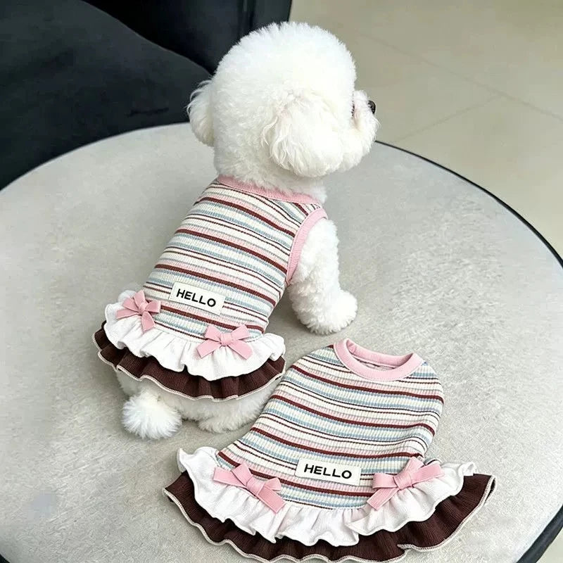 A white poodle wearing a striped dress with pink trim and ruffle details.