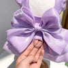 A purple satin pet dress with a large bow and a D-ring is being held by a hand.