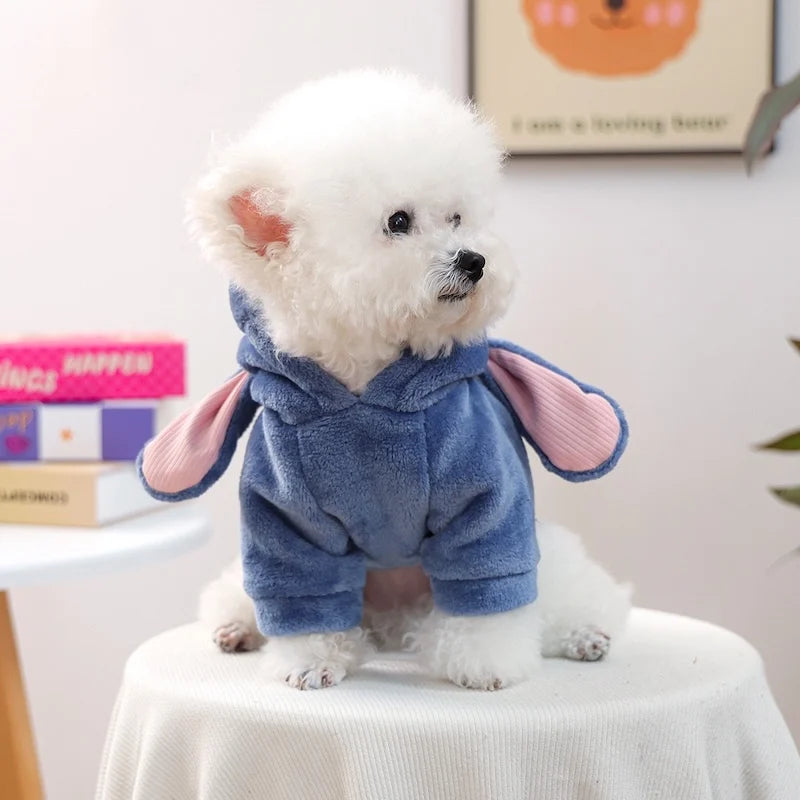 A fluffy white dog wearing a blue seasonal flannel hoodie with pink accents, sitting on a white table.