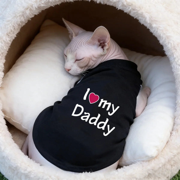 A white cat is resting inside a fluffy white pet bed wearing a black shirt with the text I love my Daddy