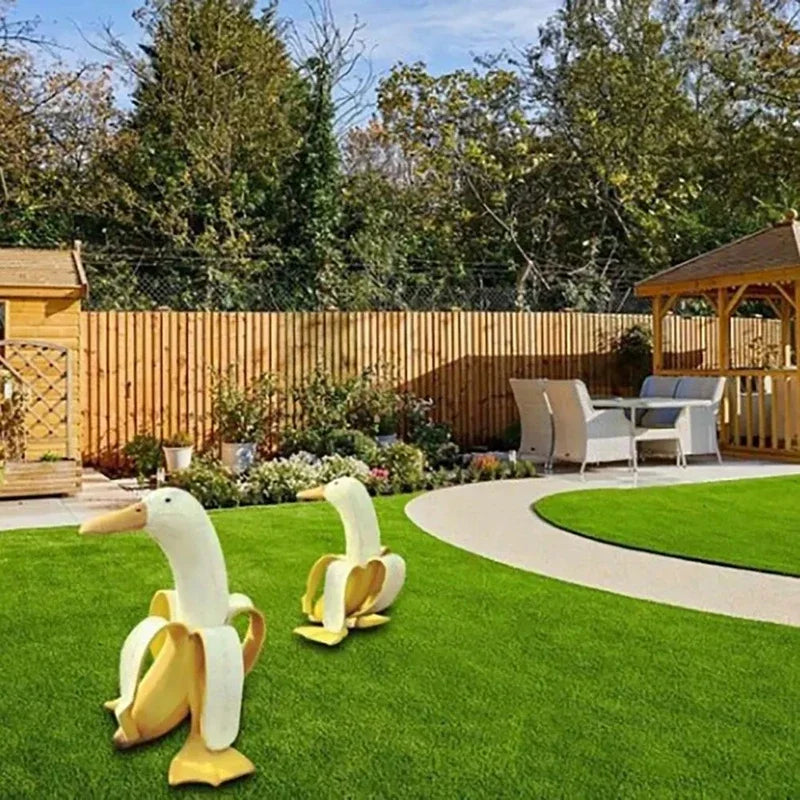 A pair of white and yellow resin banana duck garden sculptures on a green lawn near a wooden fence.