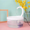 A white pet water fountain is shown pouring water into a clear bowl on a pink surface with yellow flowers in the background.