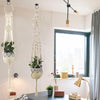 A macrame plant hanger with a leafy green plant hangs from a decorative stand in a bright room.