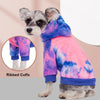 A small dog wearing a tie-dye fleece hoodie with ribbed cuffs and a purple and pink color pattern.