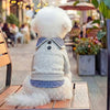 A fluffy white dog wearing a gray and blue plaid two-piece style hoodie sitting on a wooden bench outdoors.