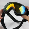 A hand is holding a pair of black adjustable dog pet goggles with yellow and blue tinted lenses.