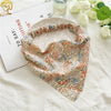 A floral paisley triangle head scarf draped on a white cloth next to a glass bottle and a white rose.