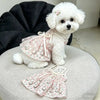 A white fluffy dog wearing a pink floral dress and a small flower accessory.