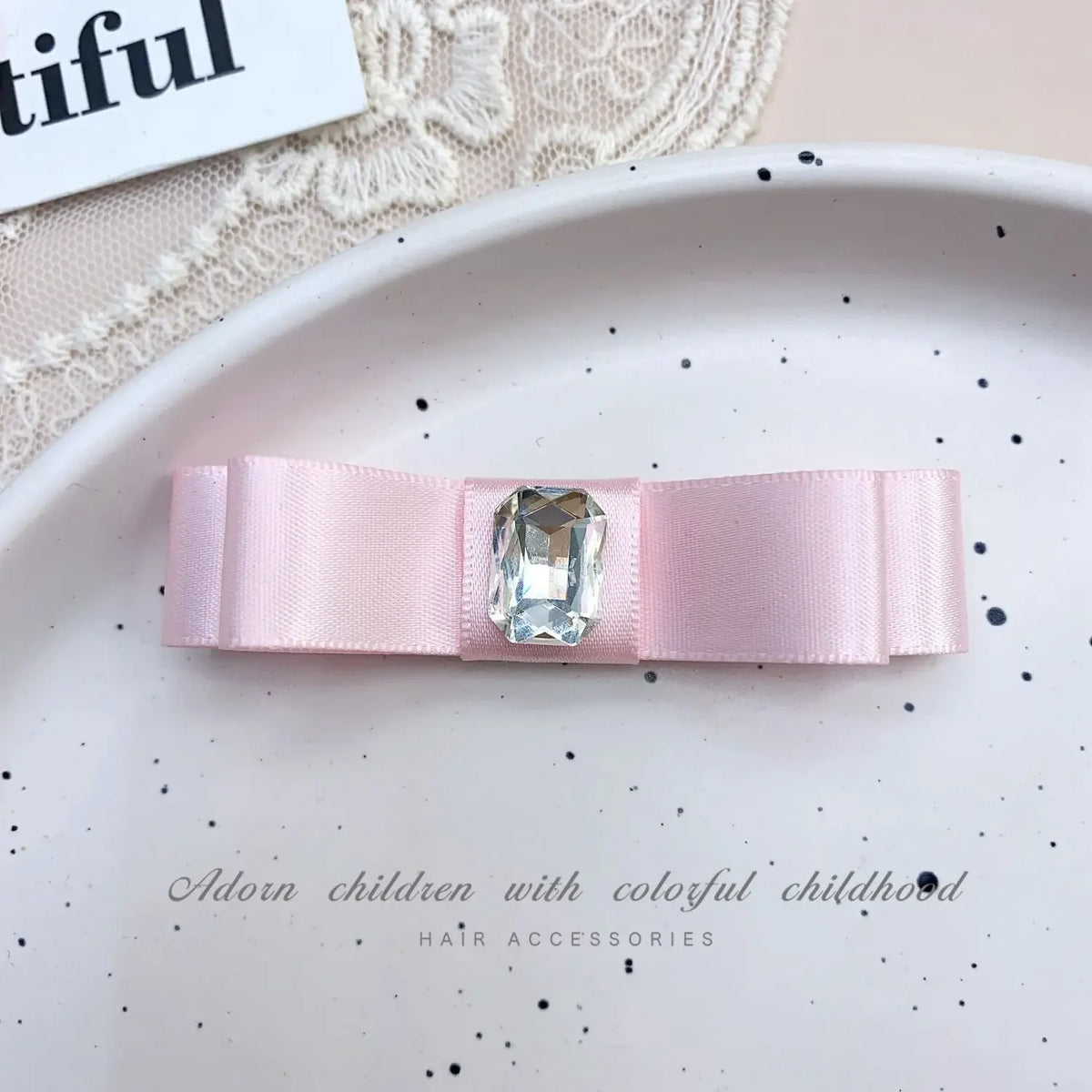 A pink satin bow hair accessory with a clear rhinestone pearl accent displayed on a white plate.