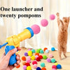 Eine Hand hält einen gelben Abschusskörper mit zwanzig bunten Pompons, mit einer Katze im Hintergrund.