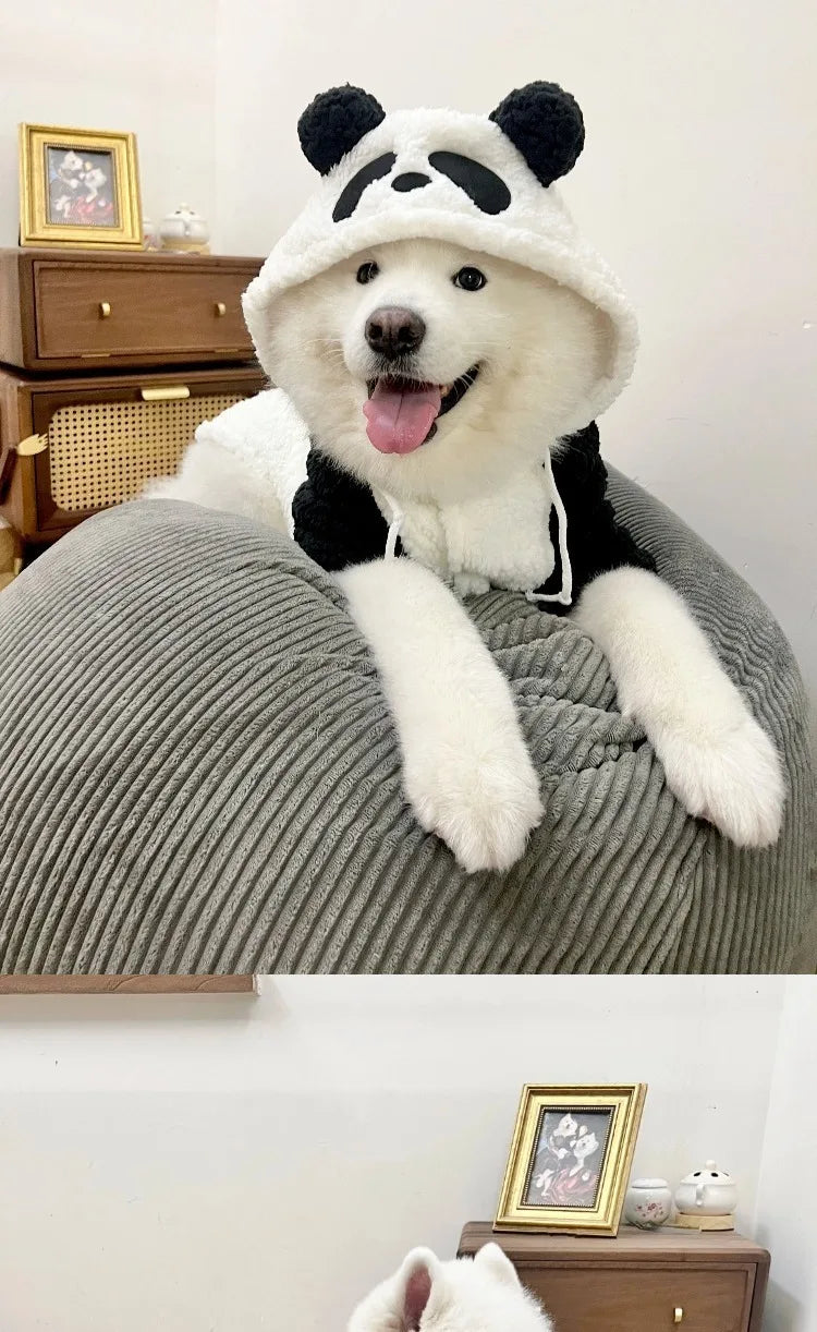 A white dog wearing a black and white panda-themed fleece hooded coat is resting on a gray fabric surface.