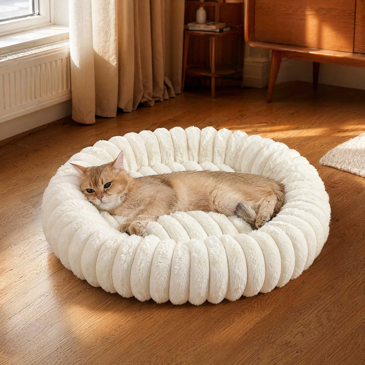 A fluffy, round pet bed with a white plush bolster is placed on a wooden floor next to a window.