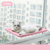 A white cat is relaxing on a pink aerial hammock pet bed indoors near a large window.
